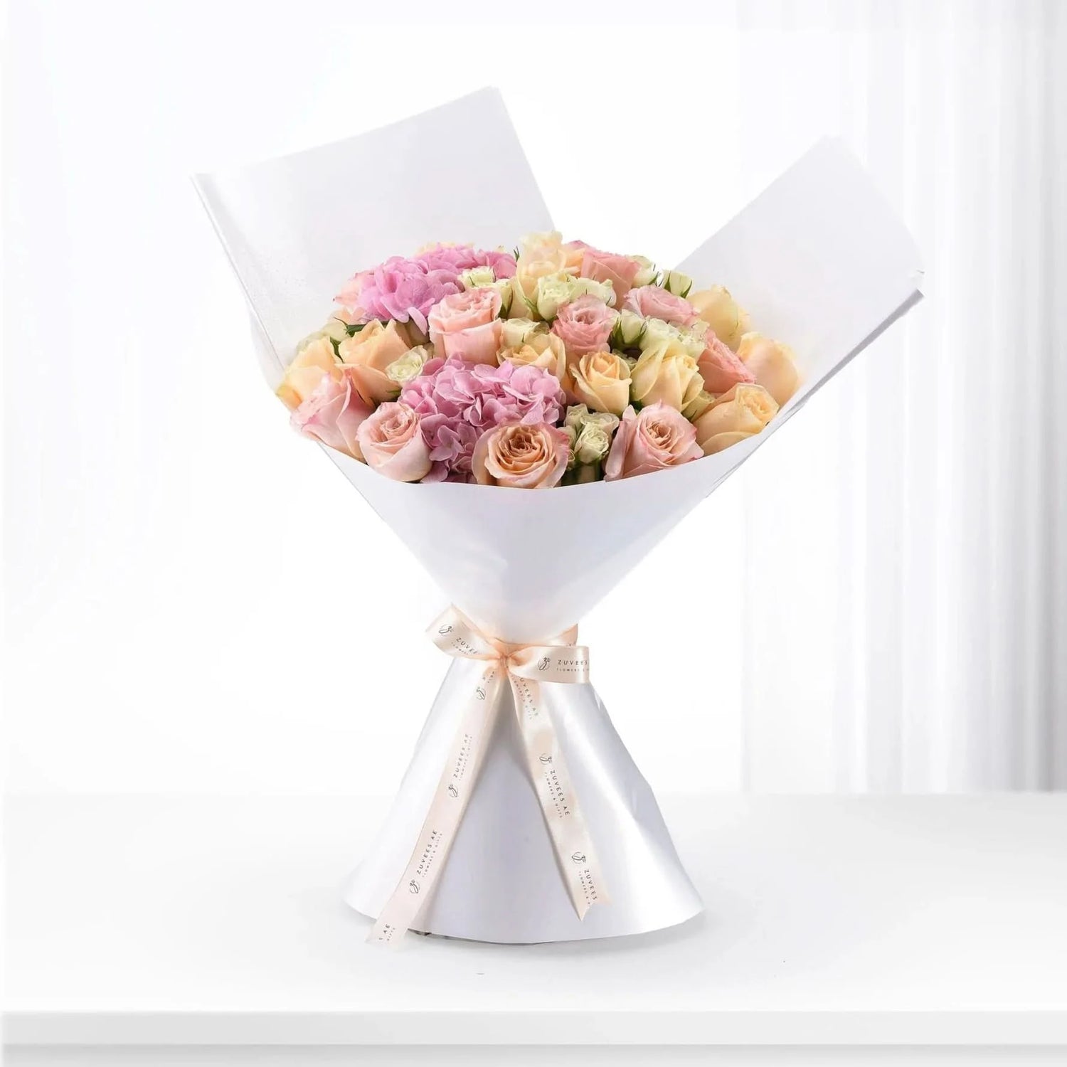 Soft pink and peach roses with hydrangeas, elegantly wrapped in white paper and ribbon.