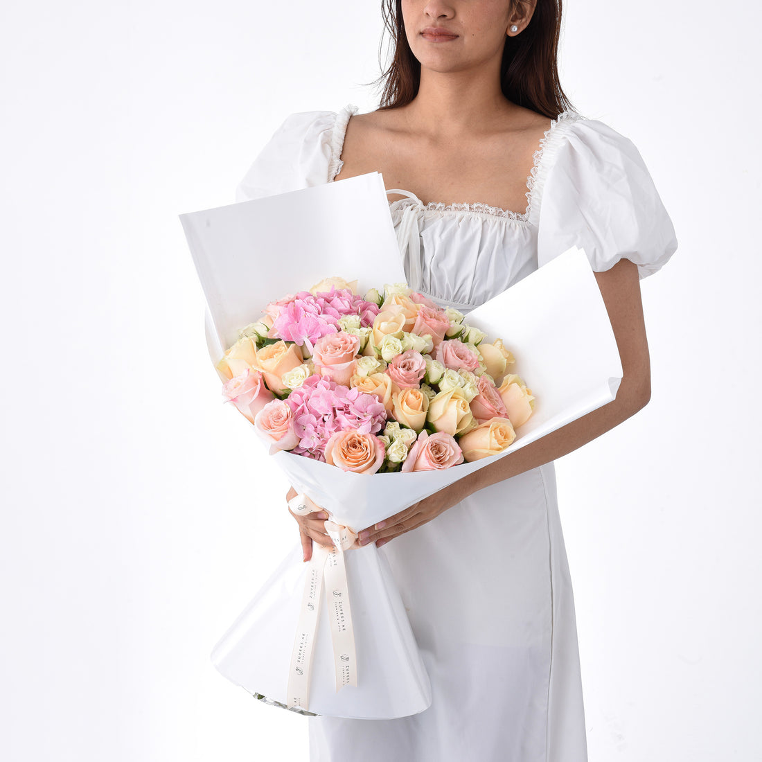 Soft pink roses and hydrangeas wrapped in crisp white paper, held by a woman in white.