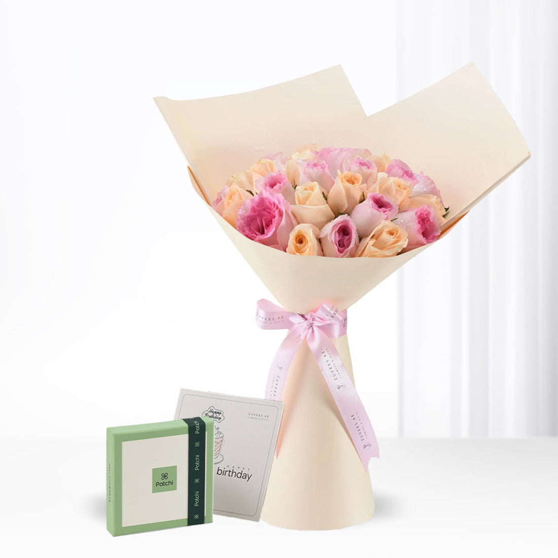 Soft pink and peach roses in cream paper, tied with pink ribbon, beside Patchi chocolate box and birthday card.