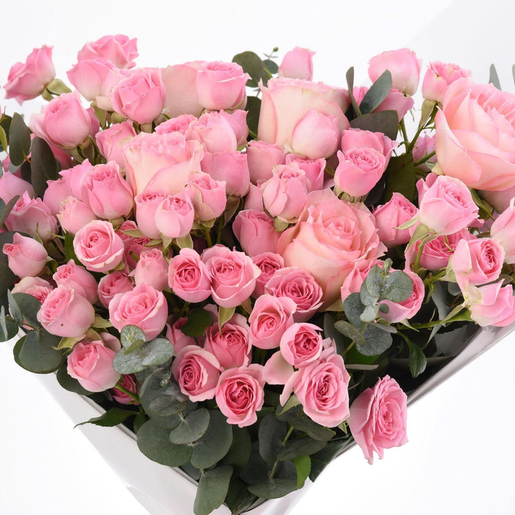 Abundant pink roses with eucalyptus in white heart-shaped box.