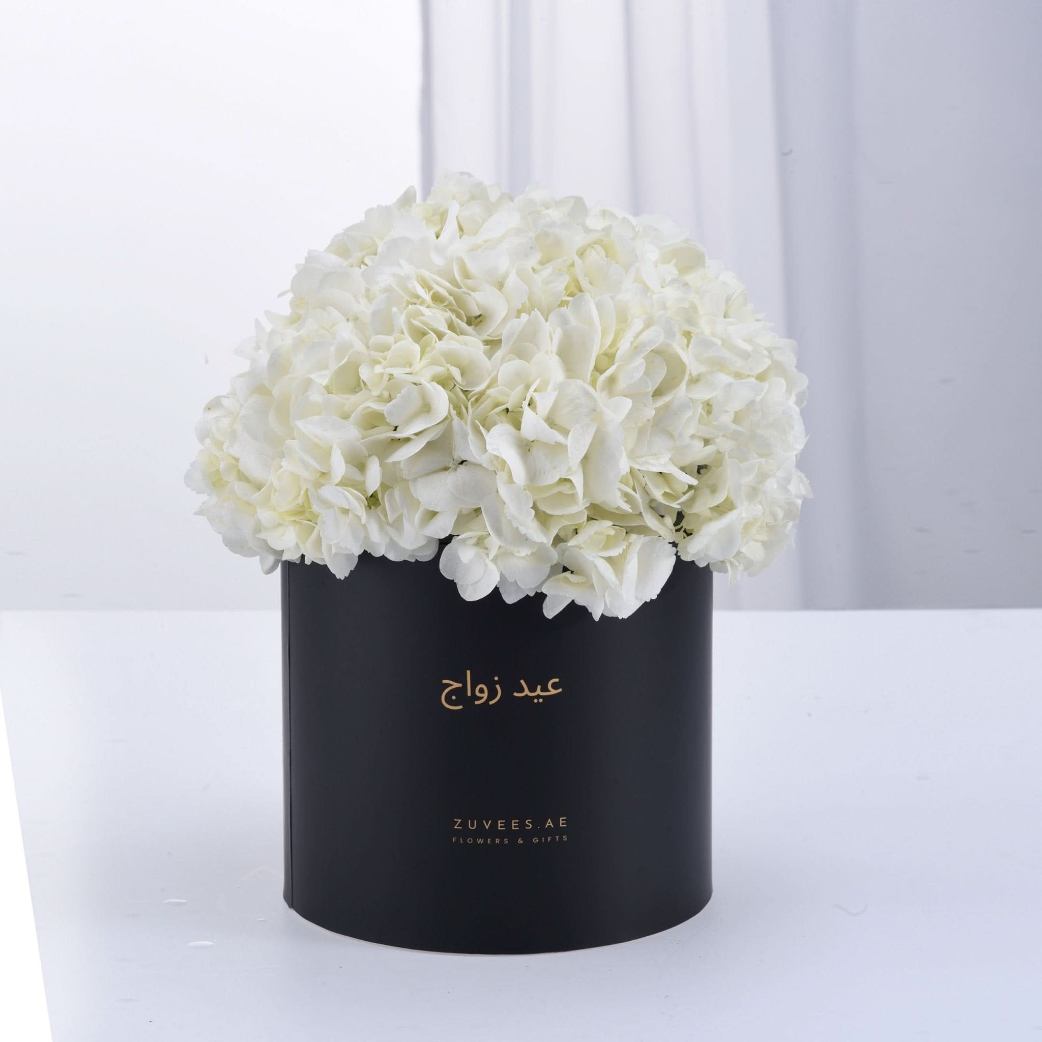 Elegant white hydrangeas in matte black box with gold Arabic script.