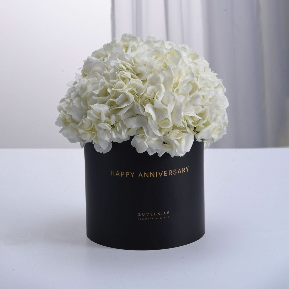 Elegant white hydrangeas in matte black box, gold "Happy Anniversary" text, minimalist luxury floral gift.