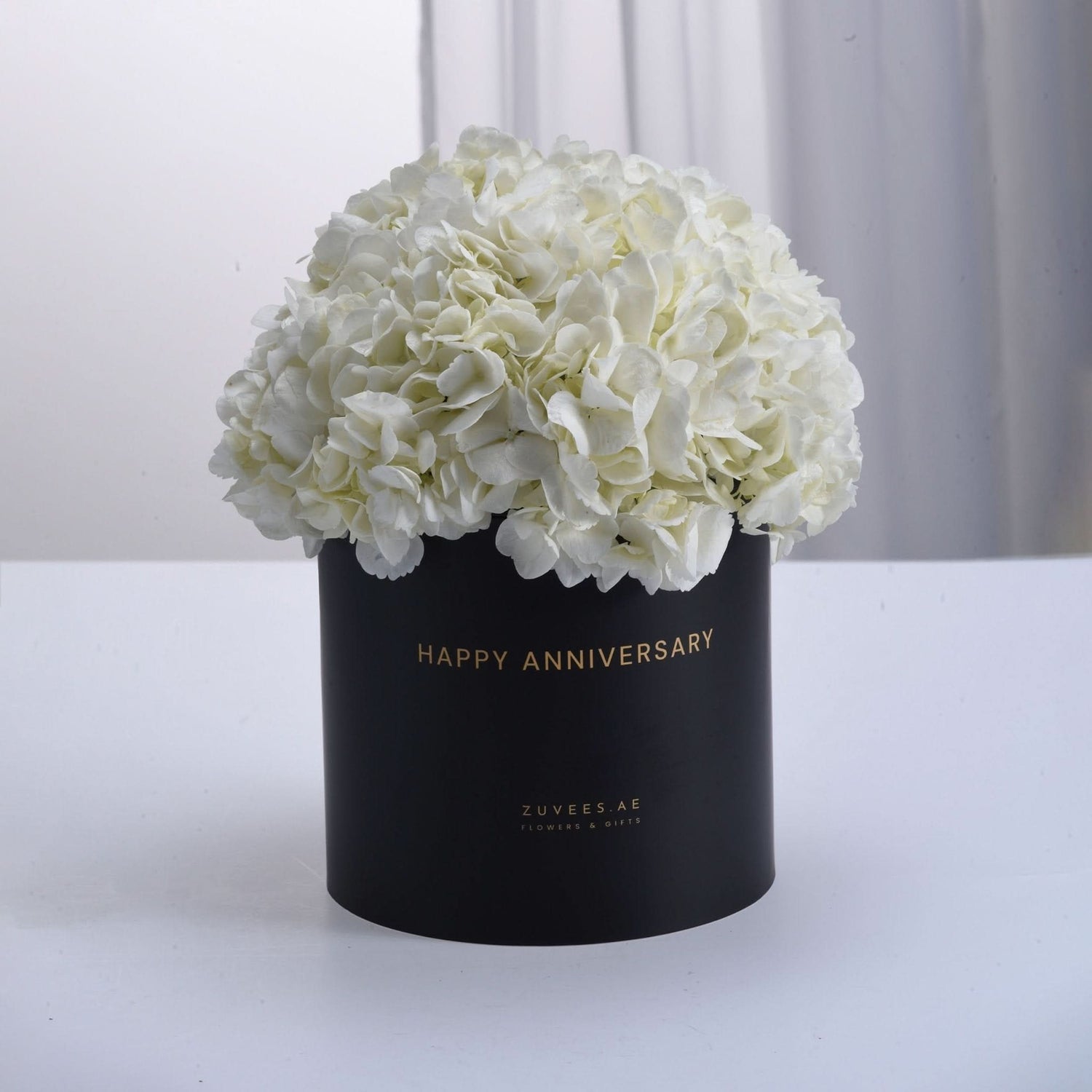Elegant white hydrangeas in matte black box, gold "Happy Anniversary" text, minimalist luxury floral gift.