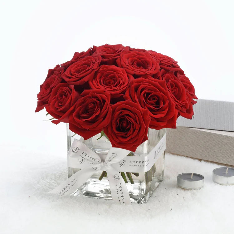 Vibrant red roses in clear glass vase with white ribbon, beside candles on soft white surface.