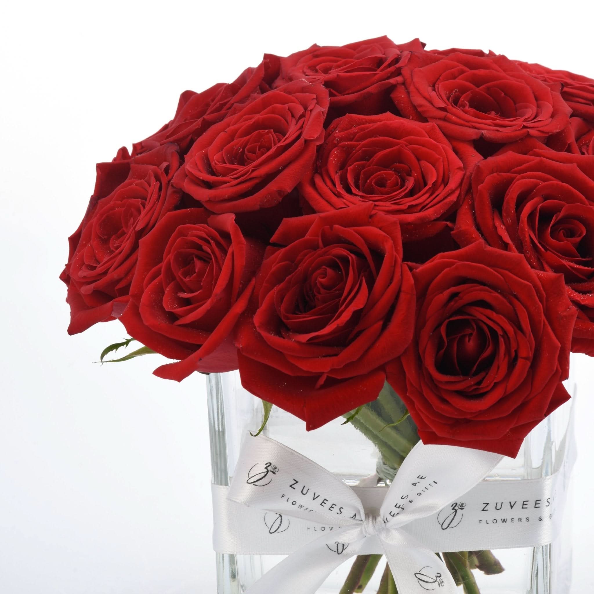 Vibrant red roses in clear glass vase, adorned with elegant white branded ribbon.