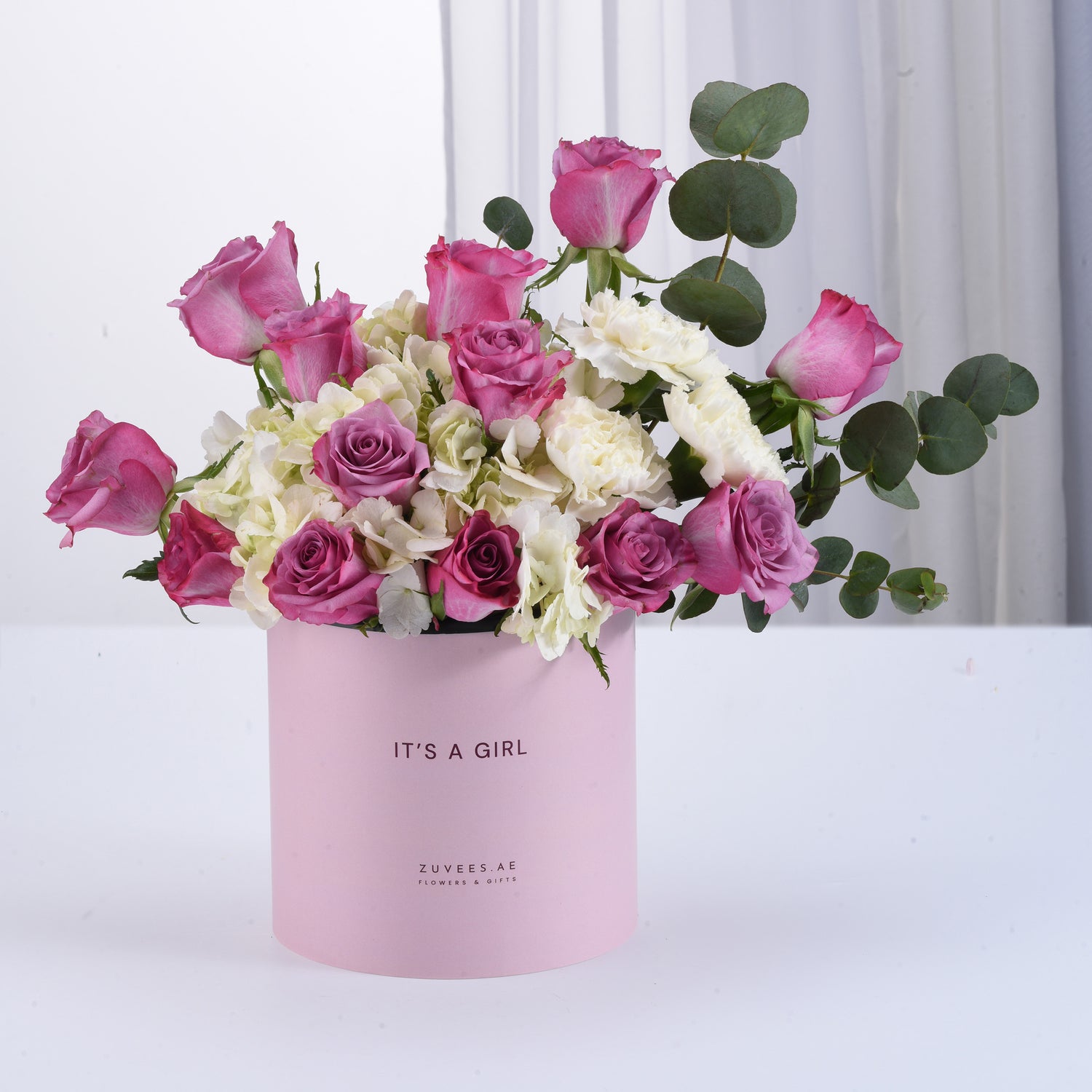 Pink box holds vibrant roses and hydrangeas, celebrating “It’s a Girl” with elegant greenery.