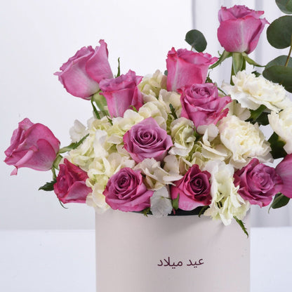 Pink roses, white hydrangeas, and eucalyptus in a cream box labeled "عيد ميلاد".