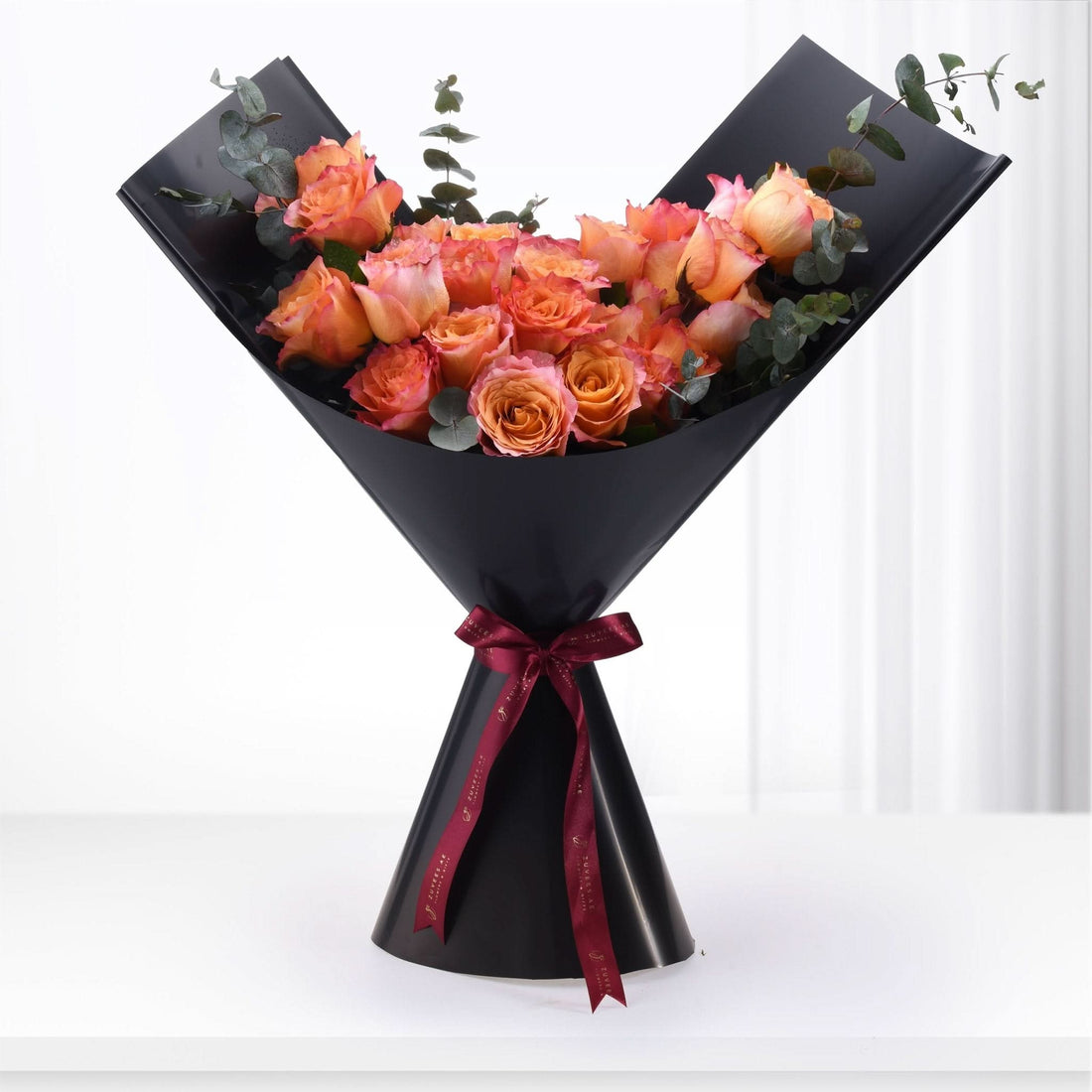 Vibrant orange-pink roses with eucalyptus, elegantly wrapped in black paper, tied with burgundy ribbon.