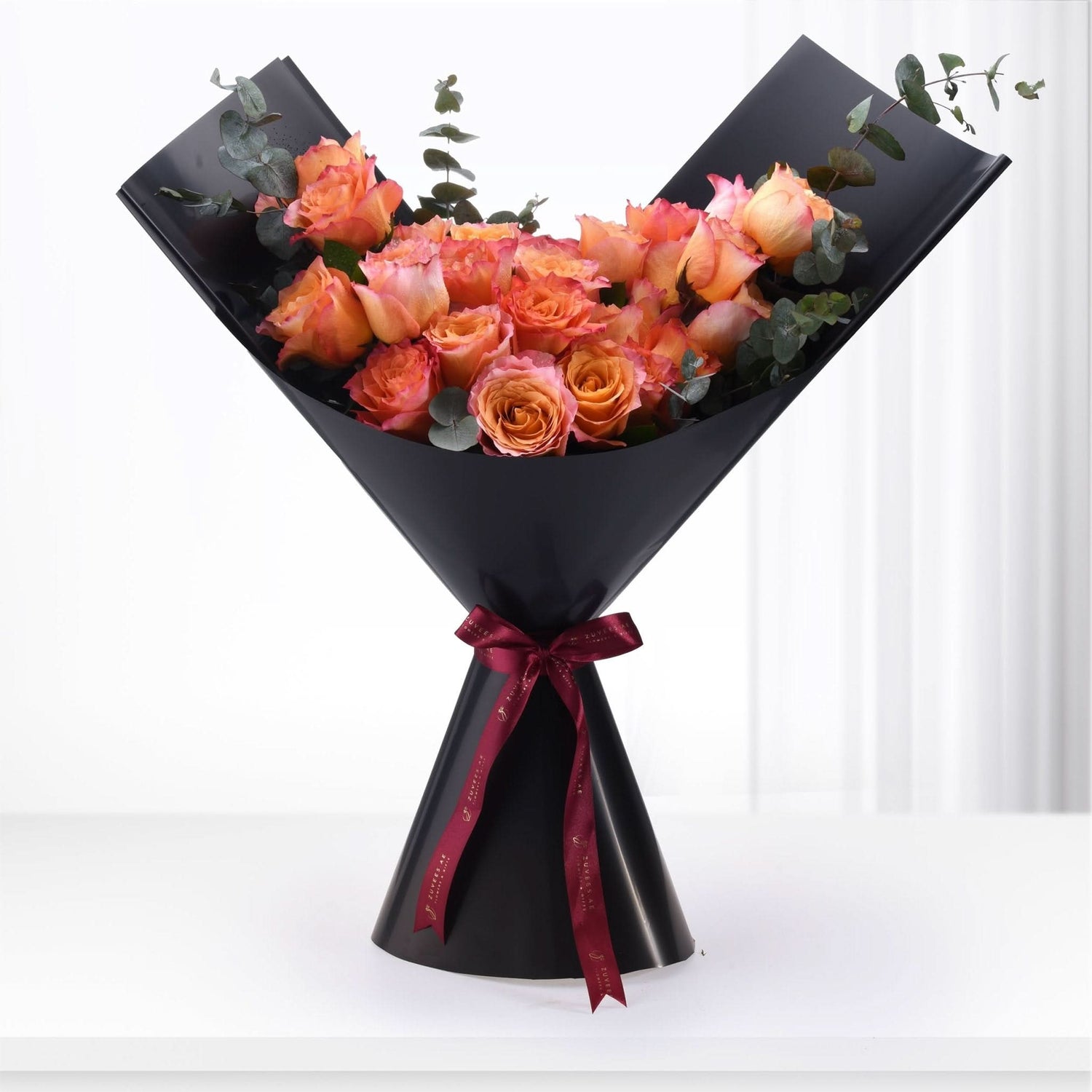 Vibrant orange-pink roses with eucalyptus, elegantly wrapped in black paper, tied with burgundy ribbon.