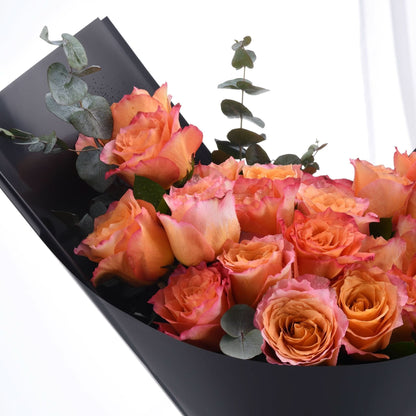 Vibrant orange-pink roses with dewdrops, accented by eucalyptus, elegantly wrapped in matte black paper.