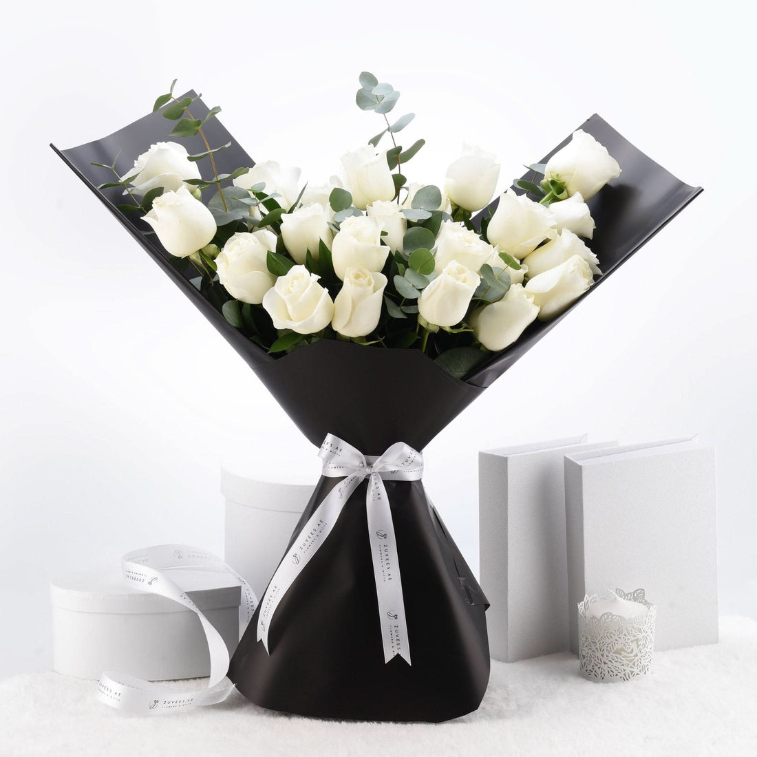 Elegant white roses in sleek black wrap, accented with silver ribbon and eucalyptus, set against minimalist white backdrop.