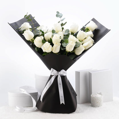 Elegant white roses in sleek black wrap, accented with silver ribbon and eucalyptus, set against minimalist white backdrop.