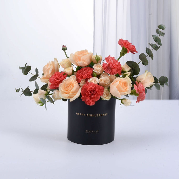 Black cylindrical box with "Happy Anniversary" text, holding peach roses and coral carnations with eucalyptus.