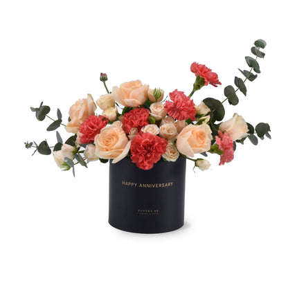 Elegant peach roses and coral carnations in sleek black "Happy Anniversary" cylinder.