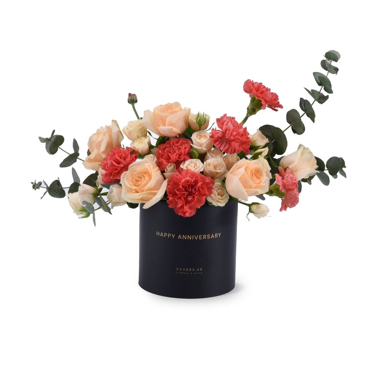 Elegant peach roses and coral carnations in sleek black "Happy Anniversary" cylinder.