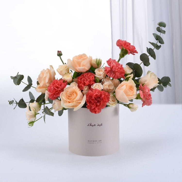 Soft peach roses and coral carnations in minimalist white box with Arabic script.
