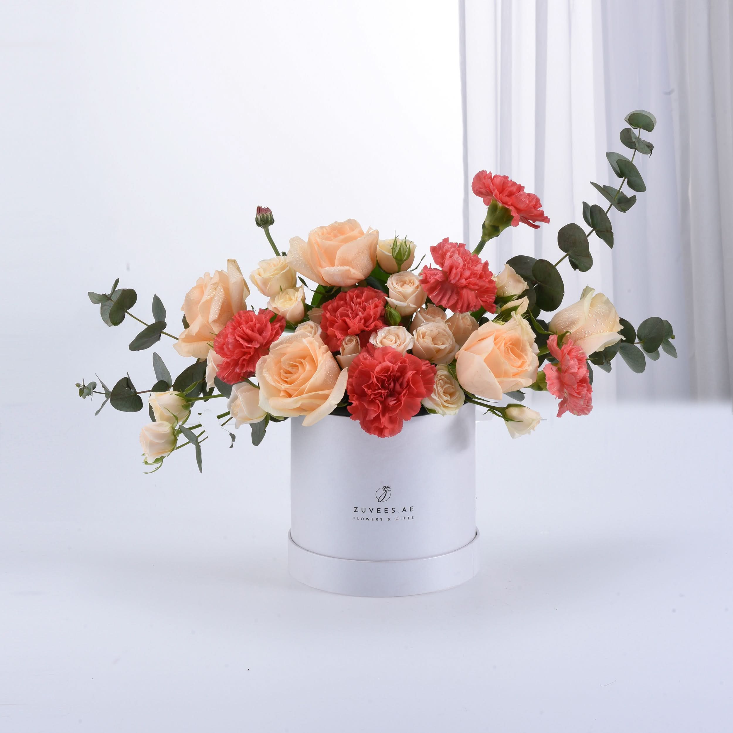 Peach roses, coral carnations, eucalyptus in white Zuvees box, dew-kissed, elegant gift arrangement.