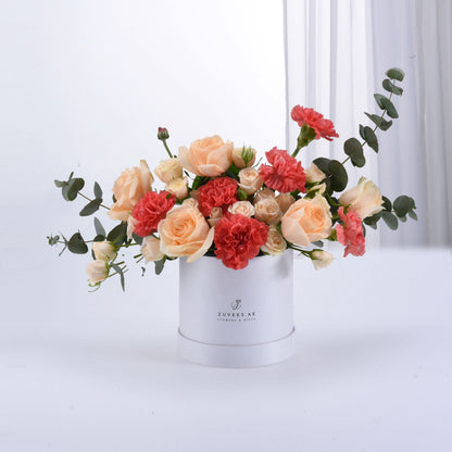Peach roses, coral carnations, eucalyptus in white Zuvees box, dew-kissed, elegant gift arrangement.