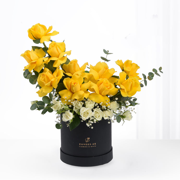 Vibrant yellow roses and white blooms elegantly arranged in a sleek black cylindrical box.