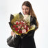 Woman holds elegant bouquet of red and pink roses with gold accents, wrapped in black and gold paper.