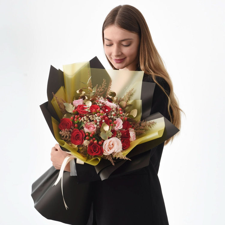 Woman holds elegant bouquet of red and pink roses with gold accents, wrapped in black and gold paper.