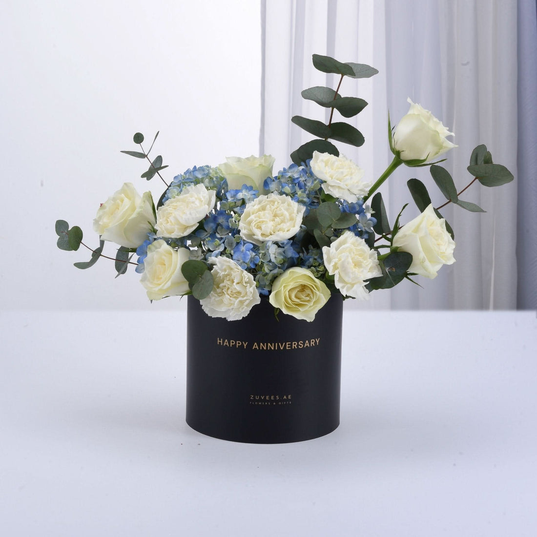 Elegant white roses, blue hydrangeas, eucalyptus in black "Happy Anniversary" box, gold lettering, minimalist white background.