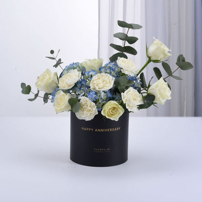Elegant white roses, blue hydrangeas, eucalyptus in black "Happy Anniversary" box, gold lettering, minimalist white background.