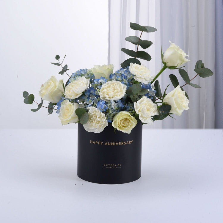 Elegant white roses, blue hydrangeas, eucalyptus in black "Happy Anniversary" box, gold lettering, minimalist white background.