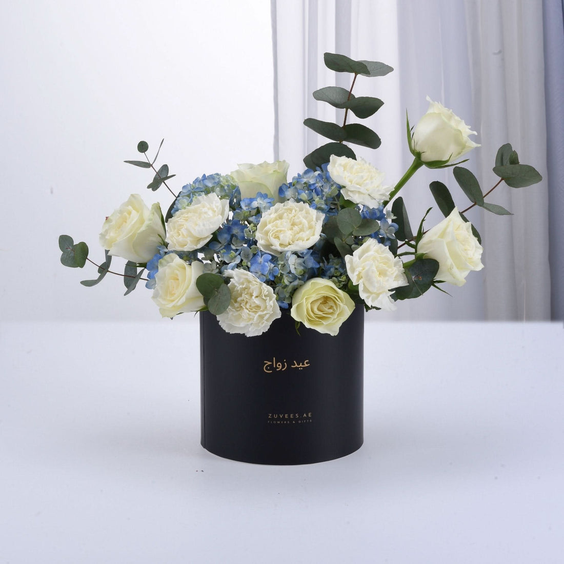 Elegant white roses and blue hydrangeas in matte black cylinder with gold Arabic script.