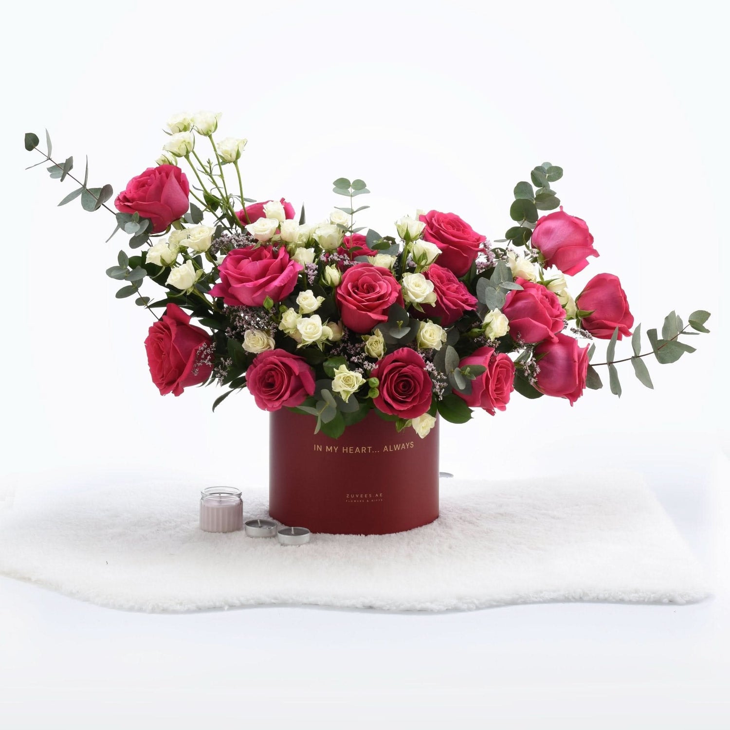 Vibrant pink and white roses with eucalyptus in deep red box, gold text, candles on white fur.