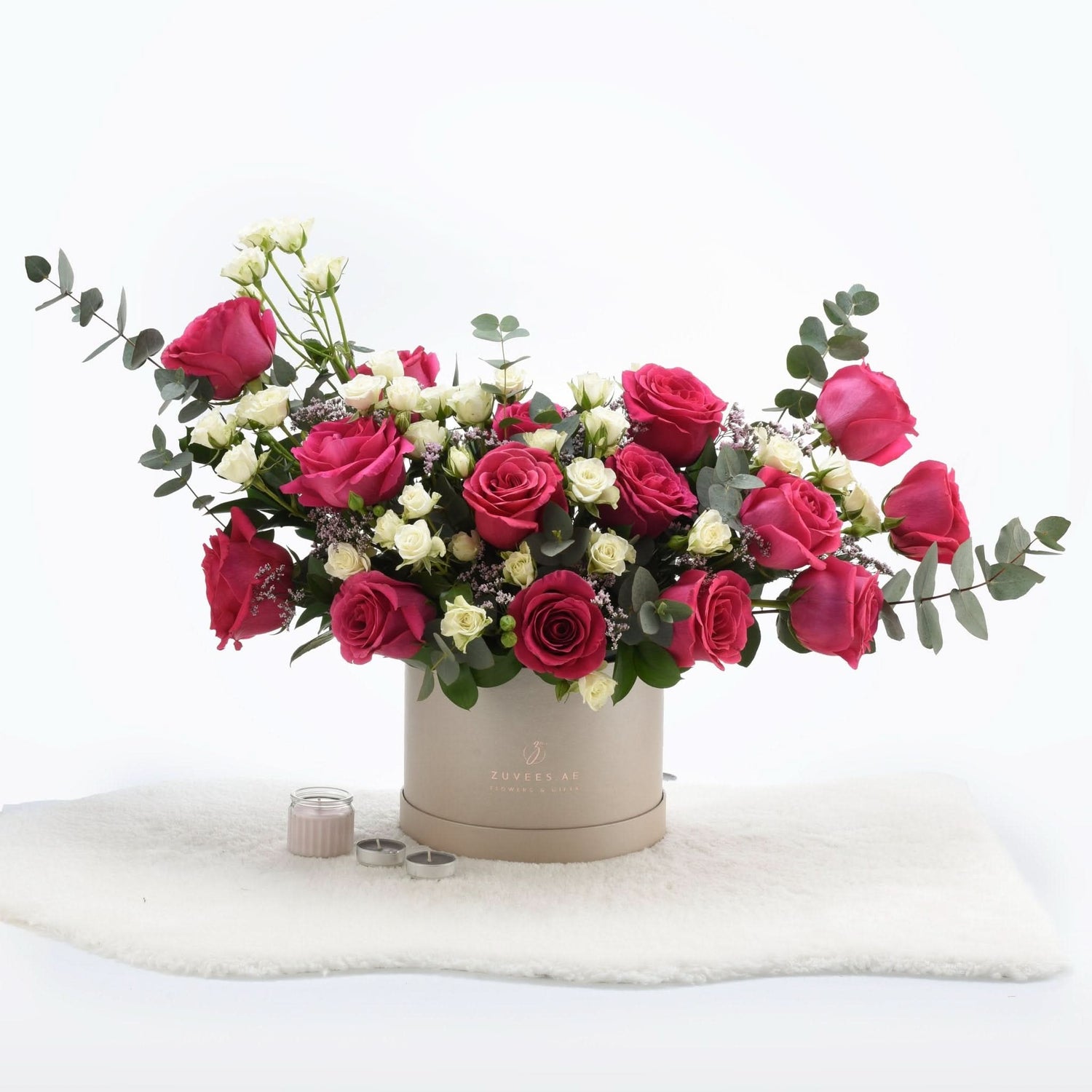Vibrant pink and white roses in beige box, accented with eucalyptus and candles.