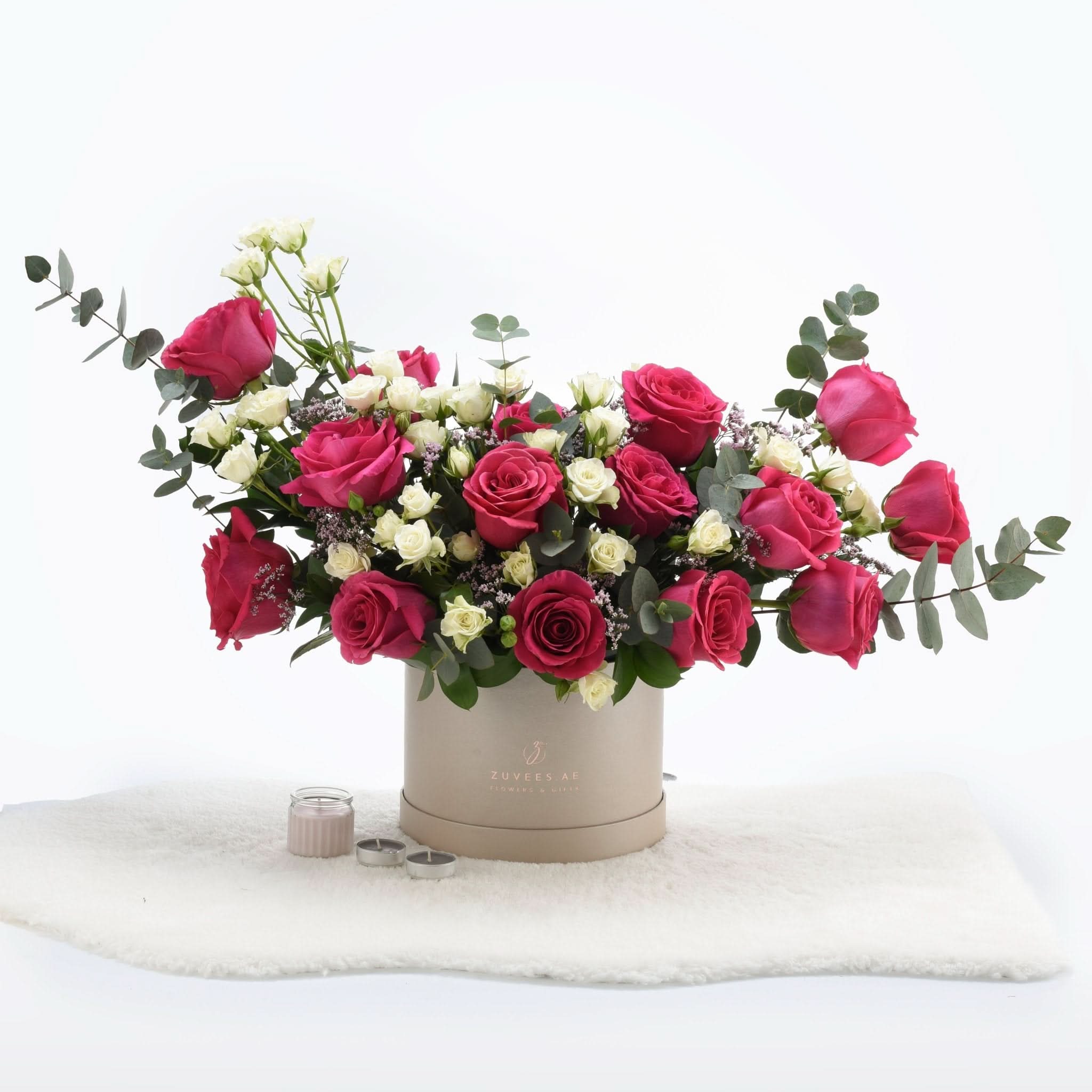 Vibrant pink and white roses in beige box, accented with eucalyptus and candles.