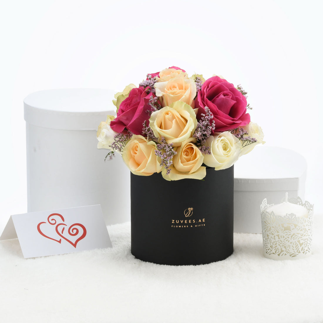Vibrant pink, peach, and cream roses in black box with gold logo, beside white candle and heart card.