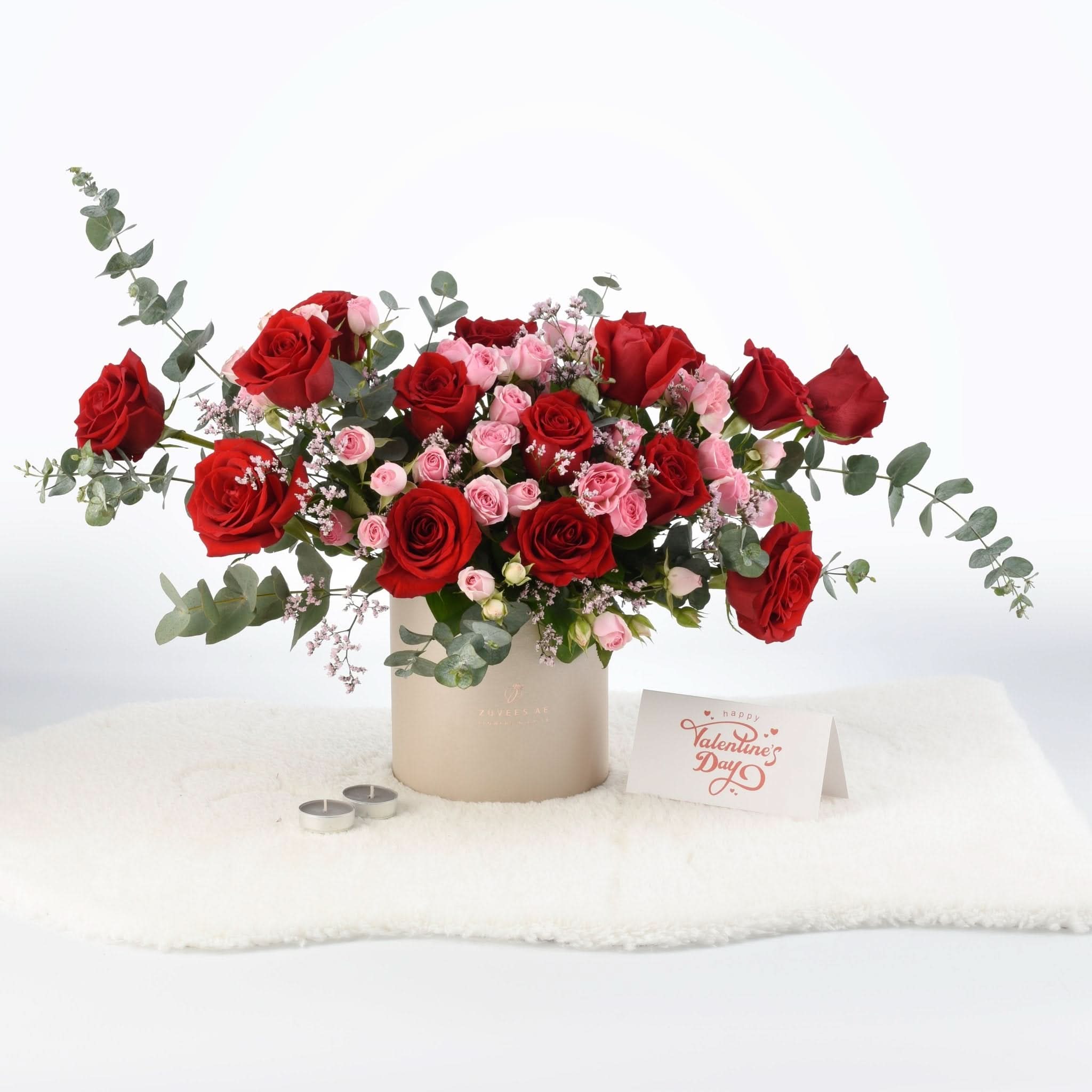 Vibrant red and pink roses in beige cylinder vase with eucalyptus, candles, and Valentine’s card.