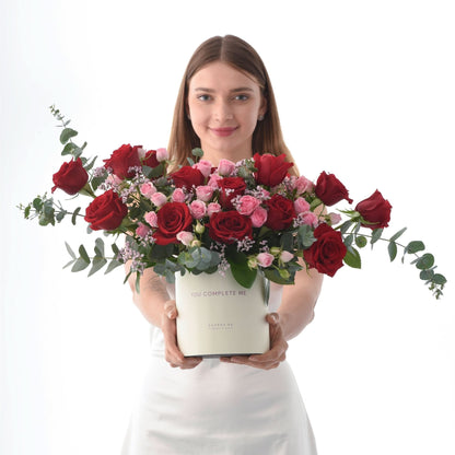 Red and pink roses with eucalyptus in cream box, held by woman in white dress.