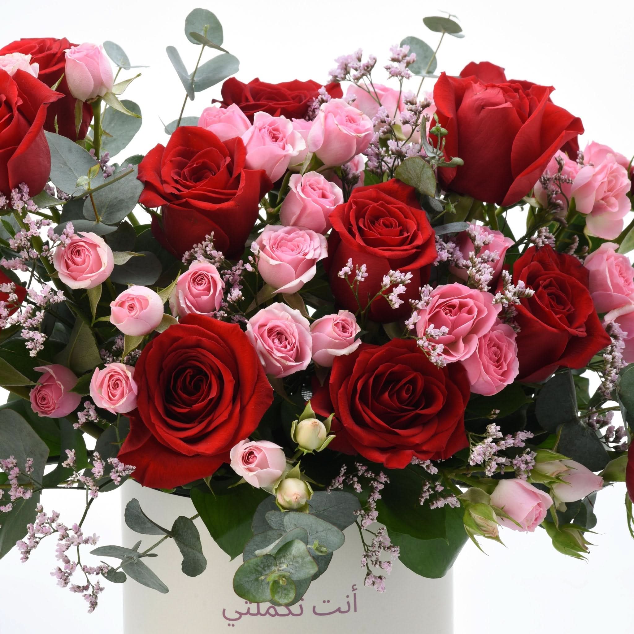 Vibrant red and pink roses with eucalyptus, in white vase with Arabic text.