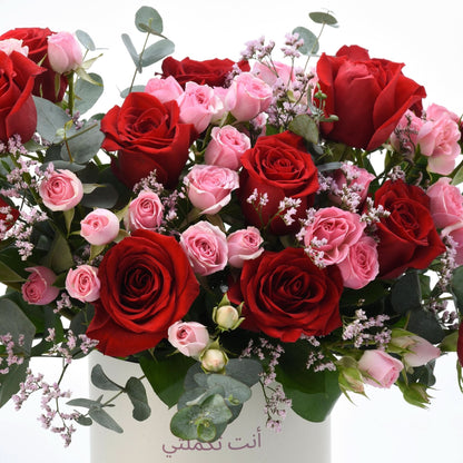 Vibrant red and pink roses with eucalyptus, in white vase with Arabic text.