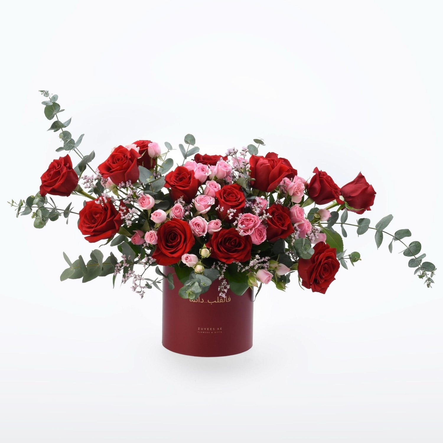 Vibrant red and pink roses cascade from a deep red cylindrical vase adorned with elegant gold text.