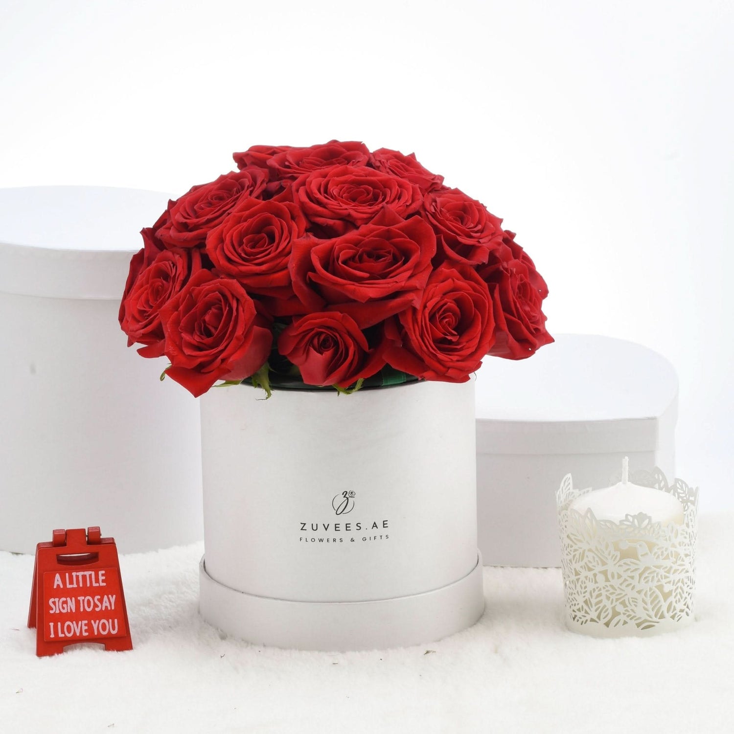 Vibrant red roses in white hat box, accented by candle and love sign.