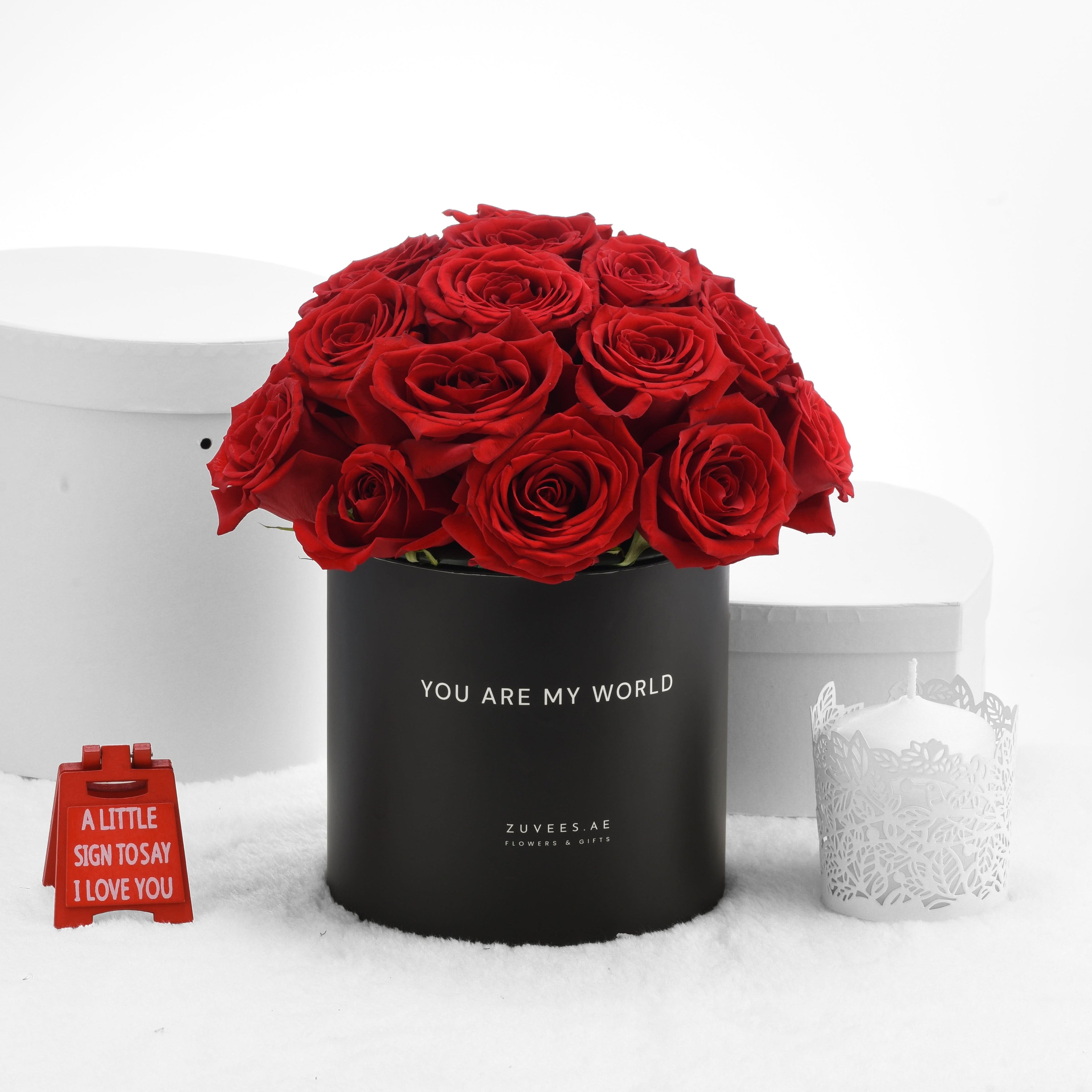 Vibrant red roses in black box with “You Are My World” text, beside white candle and red sign.