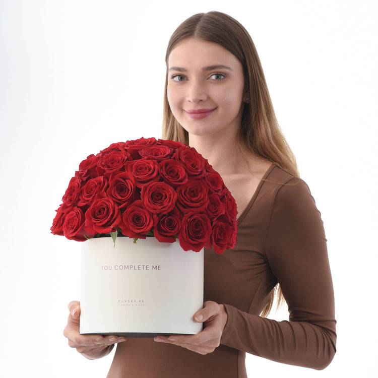 Woman holds white box with vibrant red roses, "You Complete Me" text, Zuvees.AE branding.