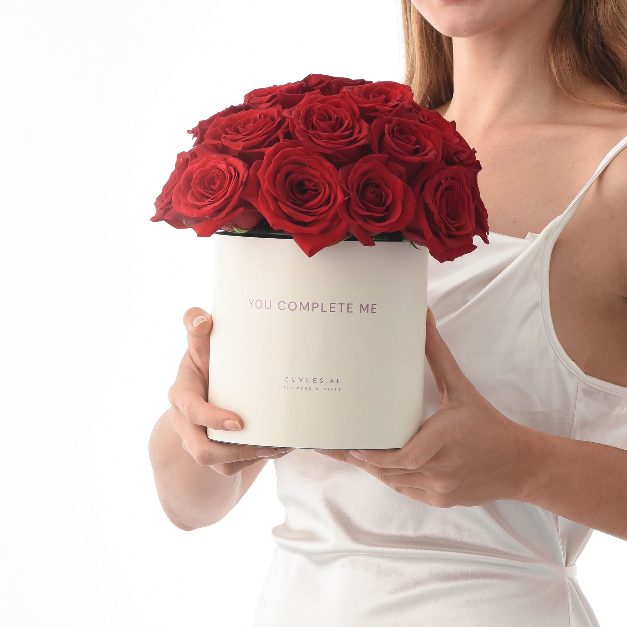 Woman holds white box of vibrant red roses with "You Complete Me" message.