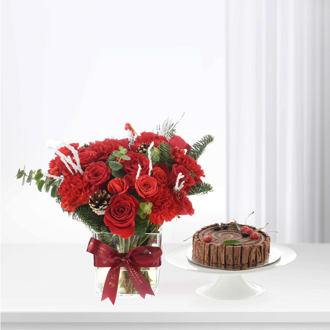 Vibrant red roses and berries in glass vase with chocolate cake on white stand.