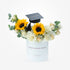 White cylindrical vase holds vibrant sunflowers, cream roses, and greenery topped with graduation cap.