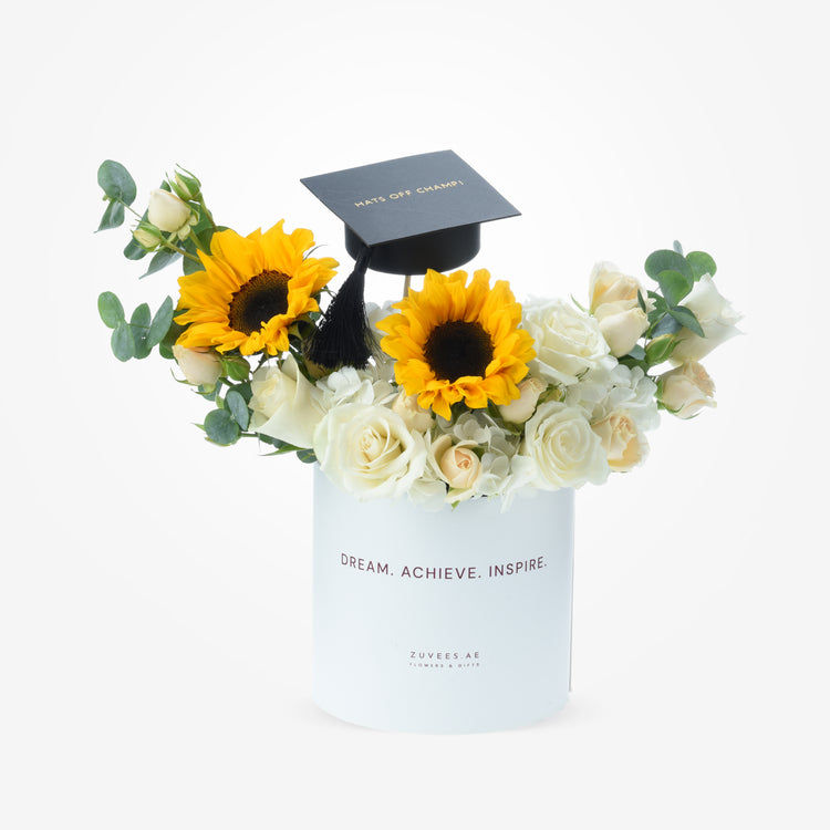 White cylindrical vase holds vibrant sunflowers, cream roses, and greenery topped with graduation cap.