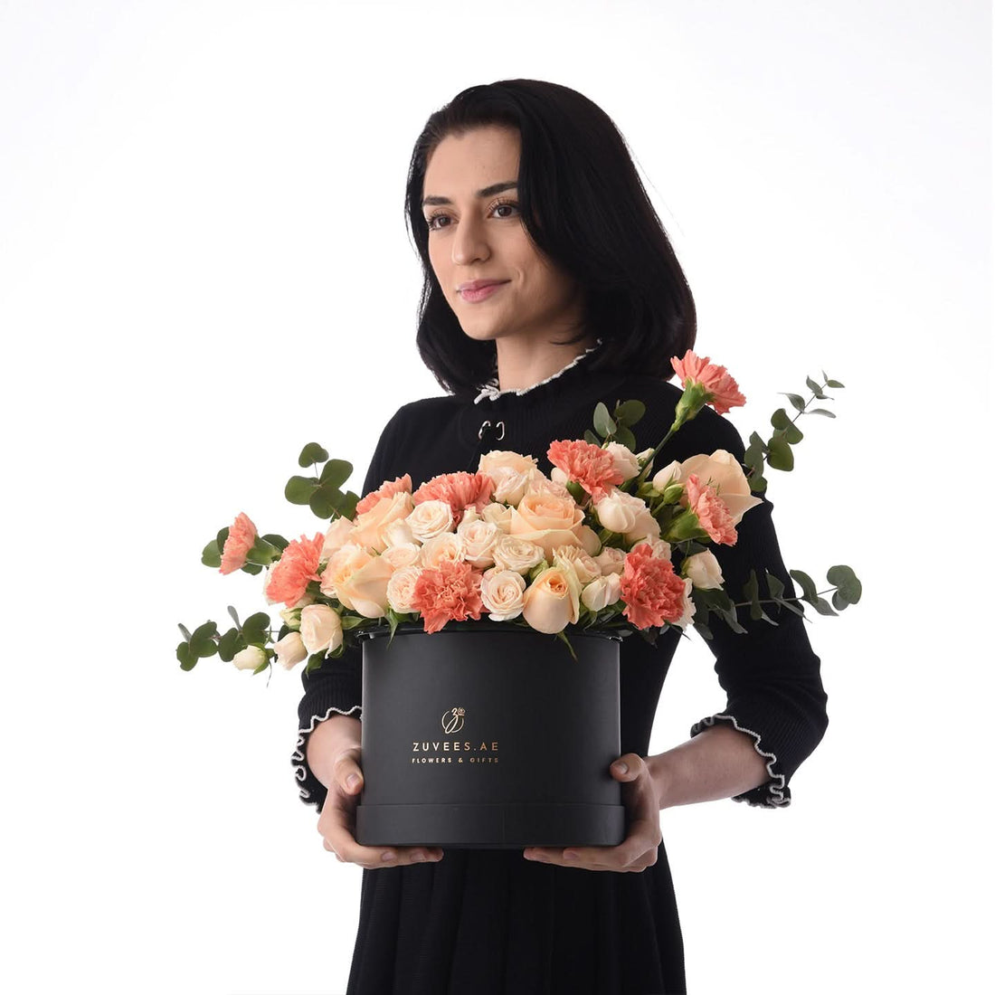 Elegant woman holds black box with peach roses and carnations against white background.
