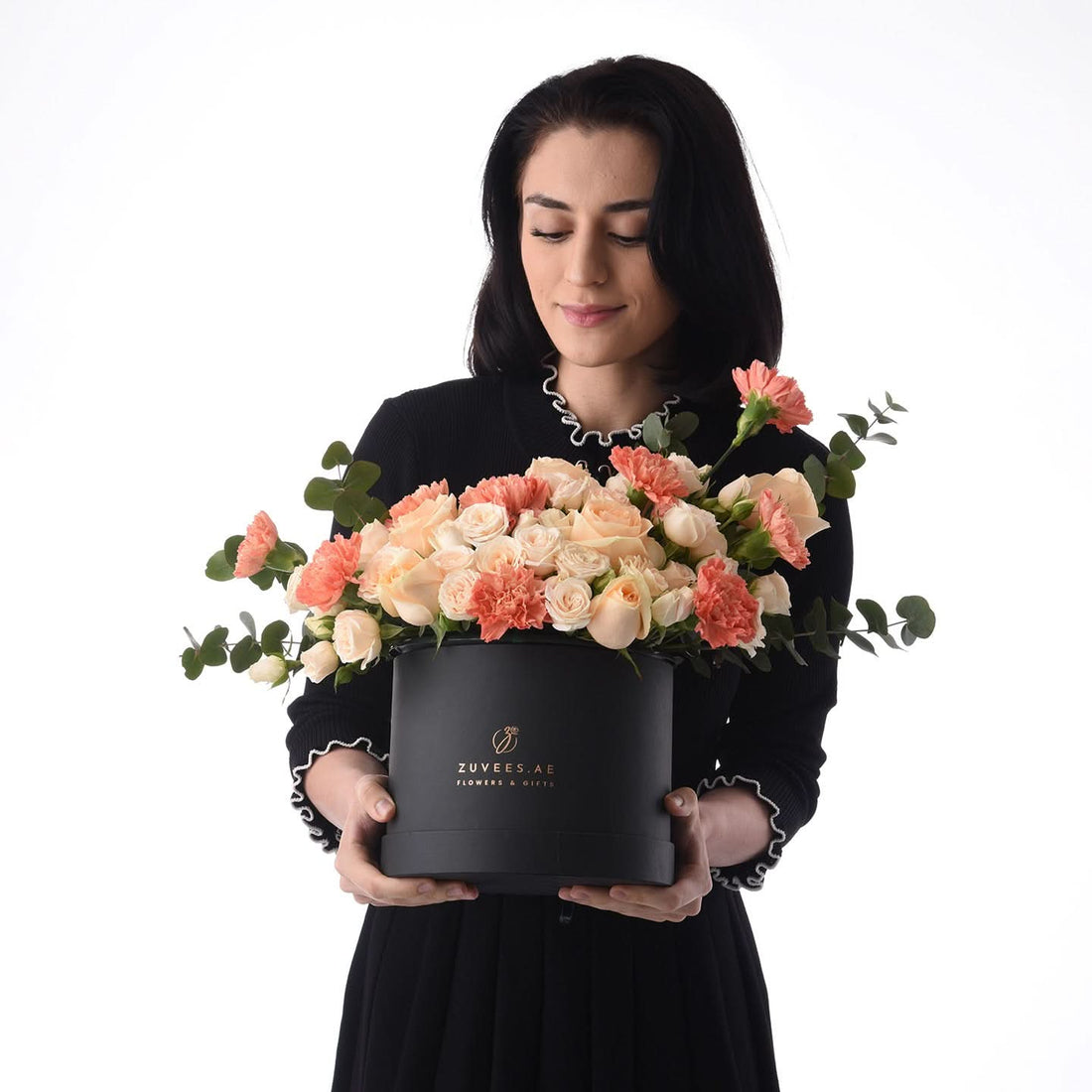 Elegant woman holds black hat box overflowing with peach roses and carnations.