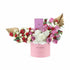 Pink cylindrical box holds vibrant red, white, and pink roses with gold leaf accents and Chanel perfume.