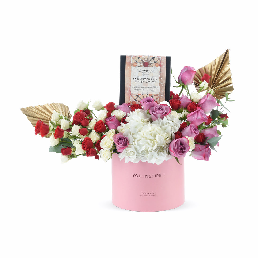 Pink cylinder holds vibrant red, white, and pink roses with white hydrangeas, accented by gold fan leaves and a "Spice Route Caramels" box.