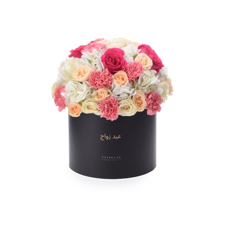 Black cylindrical box overflowing with vibrant pink, peach, and white roses and carnations.